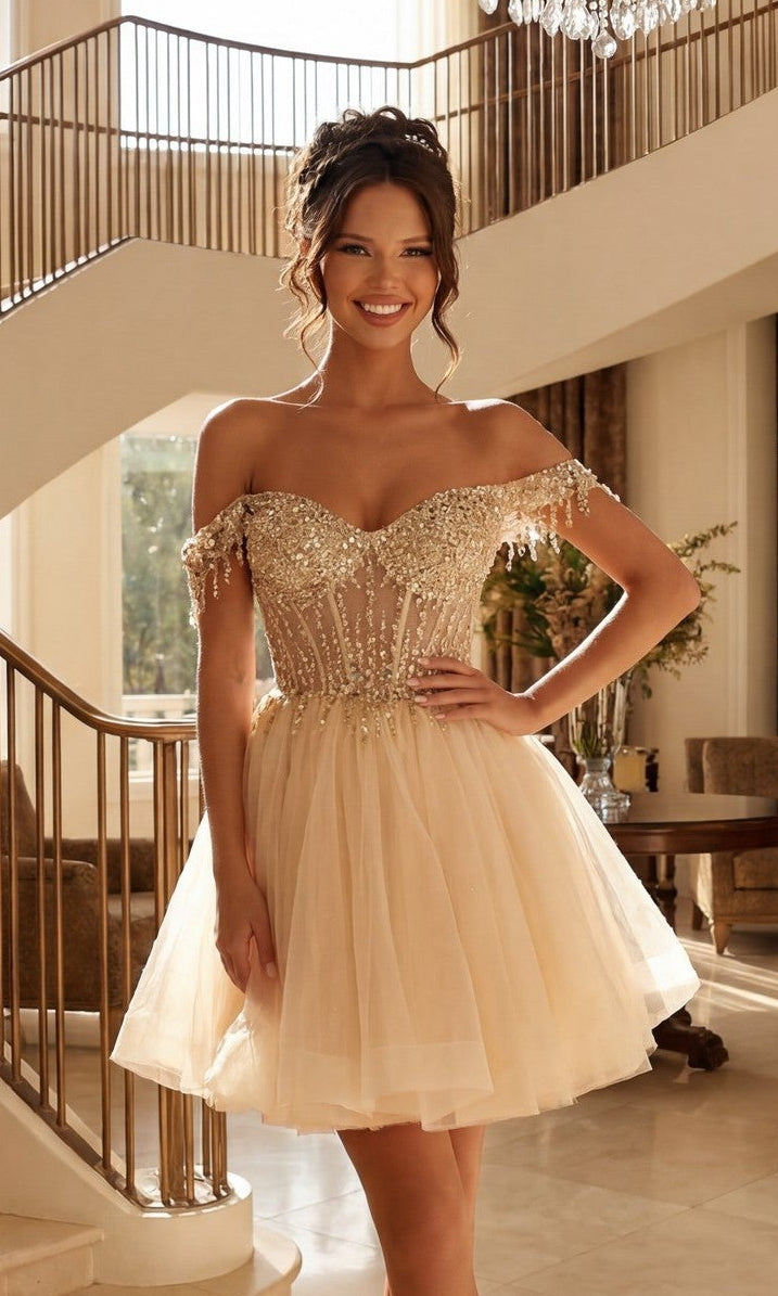 A woman stands indoors wearing the Short Champagne Corset Prom Dress: Juliet JT947L, featuring an off-the-shoulder beaded bodice and tulle skirt—a stunning champagne outfit for any party.